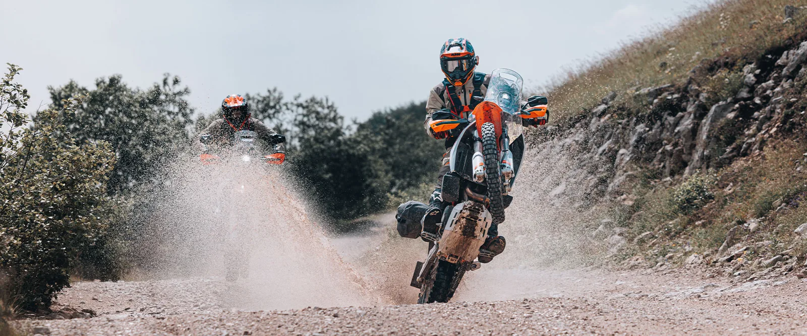 Two 2027 KTM 690 Rally motorcycles at speed on a rocky track, the lead rider standing on the pegs in a cloud of dust
