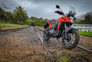 Red 2026 Suzuki V-Strom 250SX parked on a disused railway line under grey skies, showing the beak fairing, touring screen, hand guards and bash plate