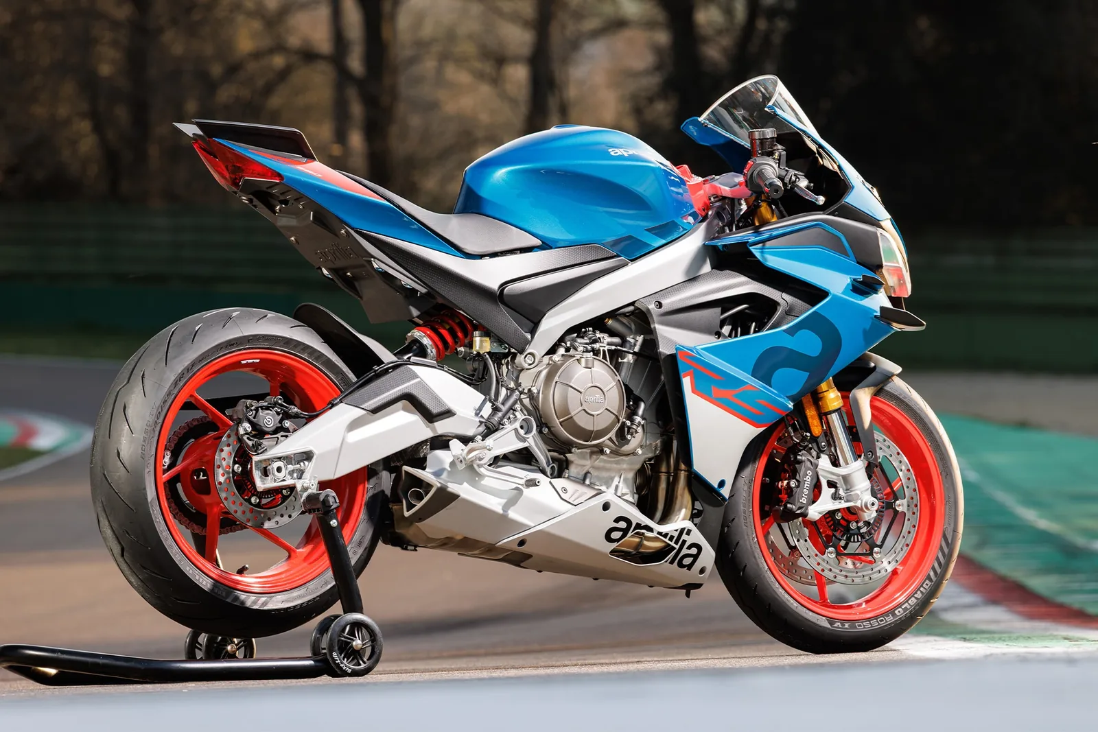 Side profile of a blue Aprilia RS 660 Factory on a paddock stand at a race track showing the Ohlins shock, Brembo calipers and red wheels