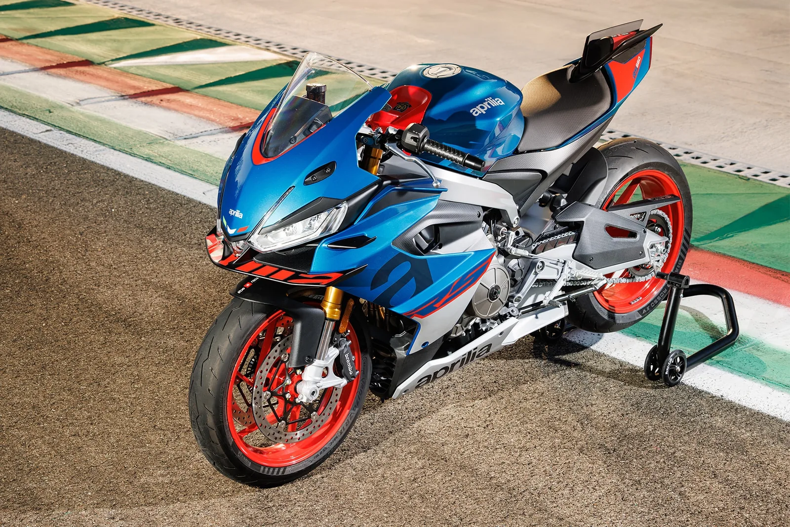 High angle view of a blue Aprilia RS 660 Factory with red wheels and gold Ohlins fork parked on a paddock stand beside a painted race-track kerb
