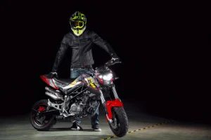 Rider in a black leather jacket and fluorescent yellow helmet standing beside a red and black Benelli TNT 125 with its four projector headlights lit in a dark studio