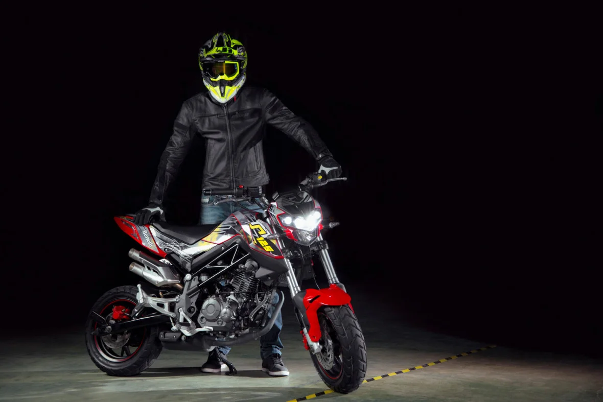 Rider in a black leather jacket and fluorescent yellow helmet standing beside a red and black Benelli TNT 125 with its four projector headlights lit in a dark studio