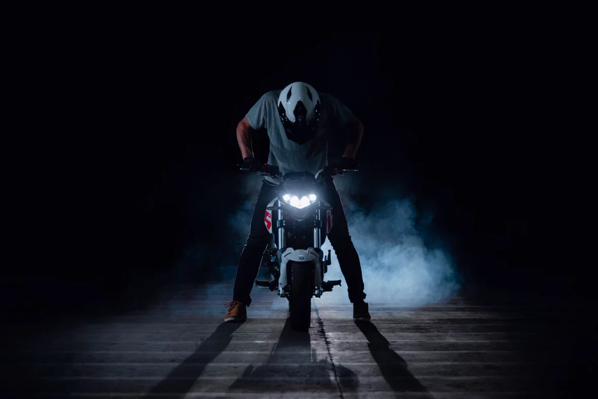 Rider in a white helmet crouched over a white Benelli TNT 125 with its headlights on in a dark smoky studio