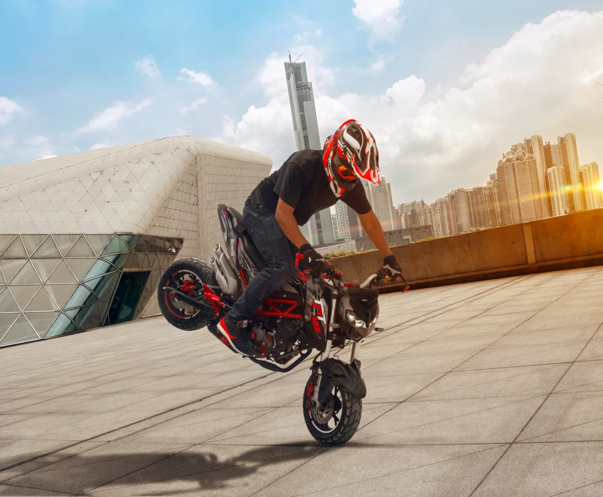 Rider in a red and white helmet performing a stoppie on a red Benelli TNT 125 on a rooftop with a city skyline behind