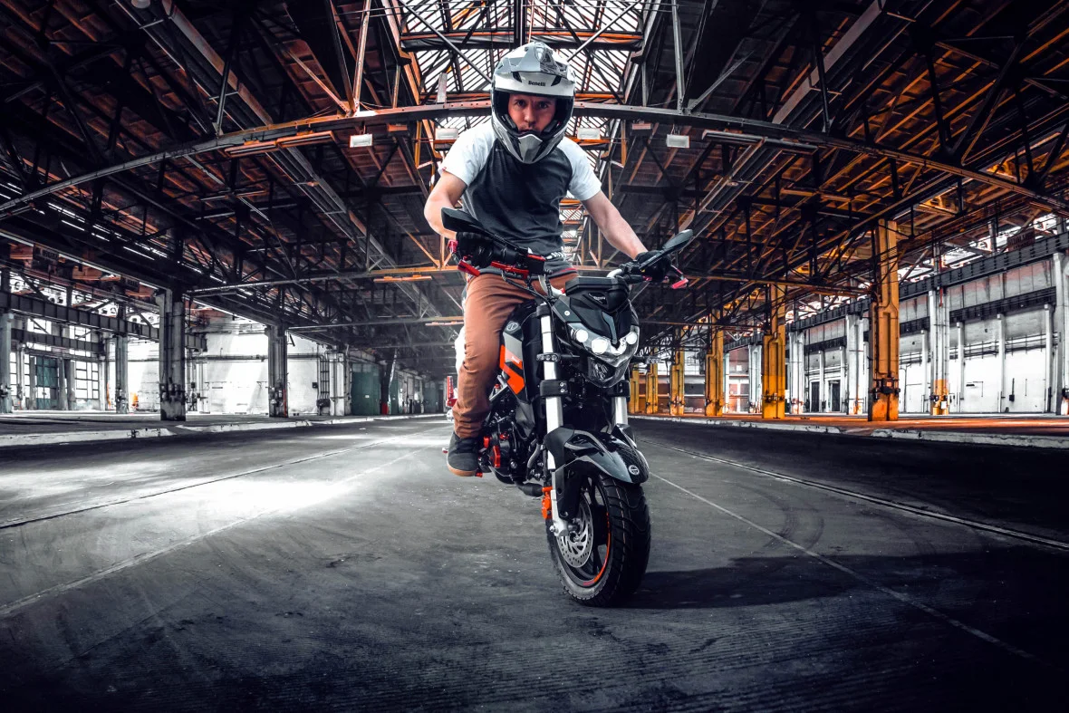 Rider in a white helmet pulling a wheelie on an orange and black Benelli TNT 125 inside an empty industrial warehouse