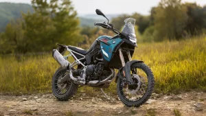 BMW F 900 GS in blue and black parked on a rocky dirt track in long grass with trees behind, front three-quarter view showing the Akrapovic silencer, gold Showa fork and Karoo 4 tyres