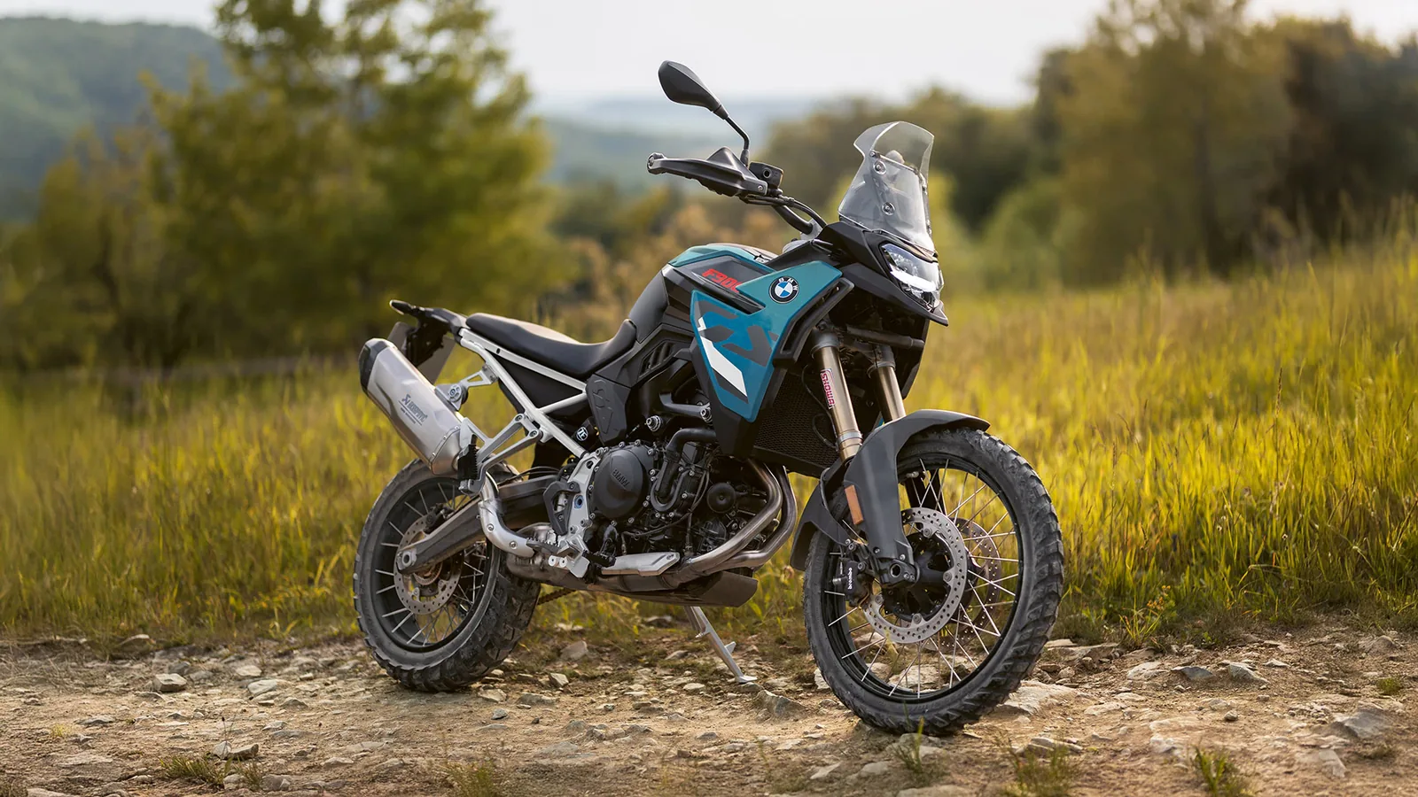 BMW F 900 GS in blue and black parked on a rocky dirt track in long grass with trees behind, front three-quarter view showing the Akrapovic silencer, gold Showa fork and Karoo 4 tyres