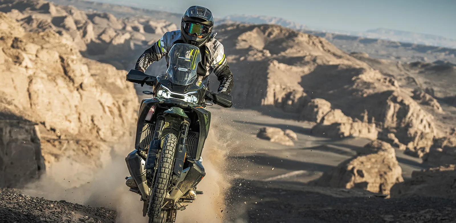 Tactical Green CFMoto 1000MT-X ridden towards the camera up a gravel track above a rocky canyon, front wheel light over a crest