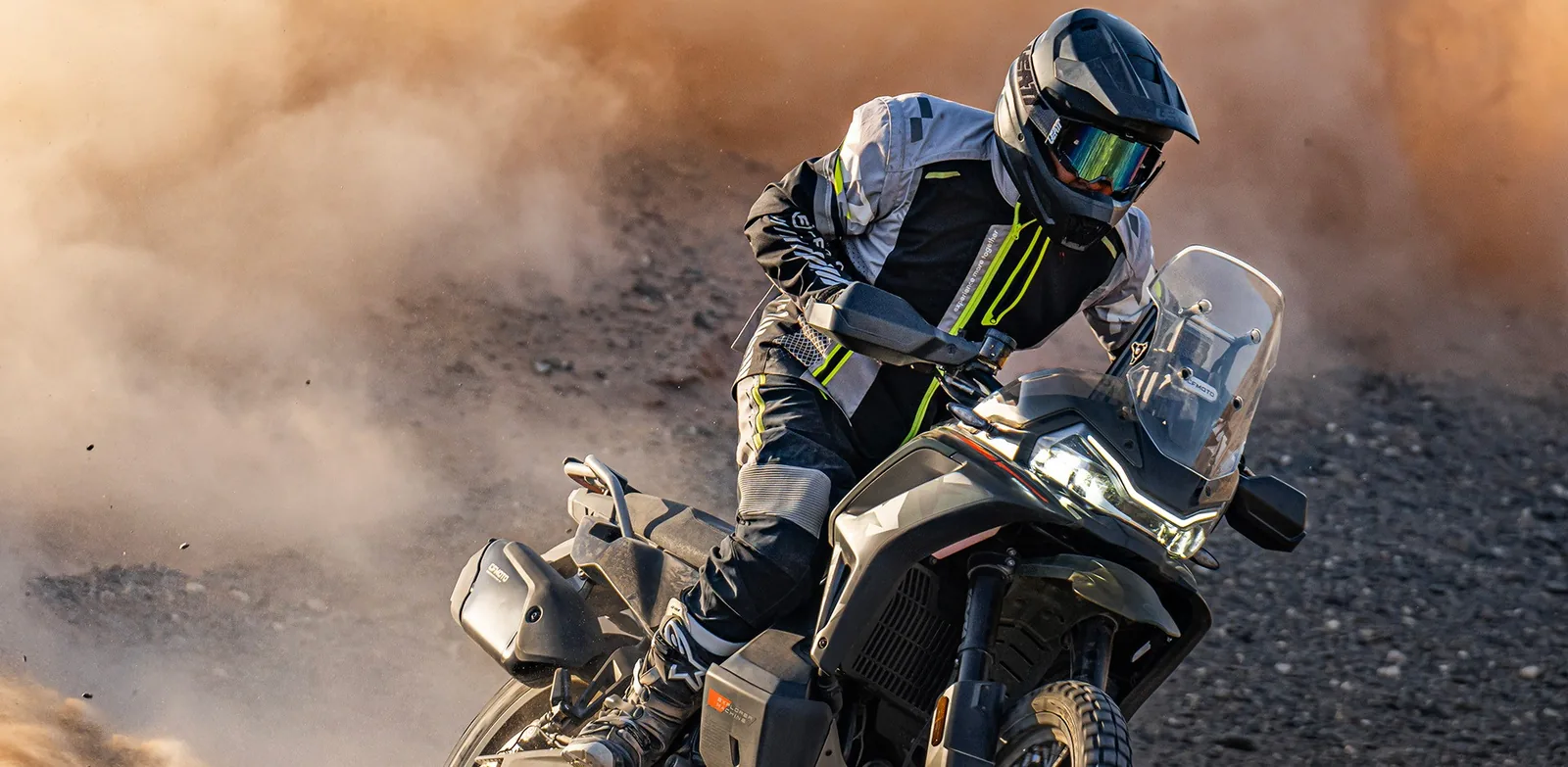 Close view of a rider cornering a Tactical Green CFMoto 1000MT-X in a cloud of orange dust, showing the LED headlight, windscreen and hand guards