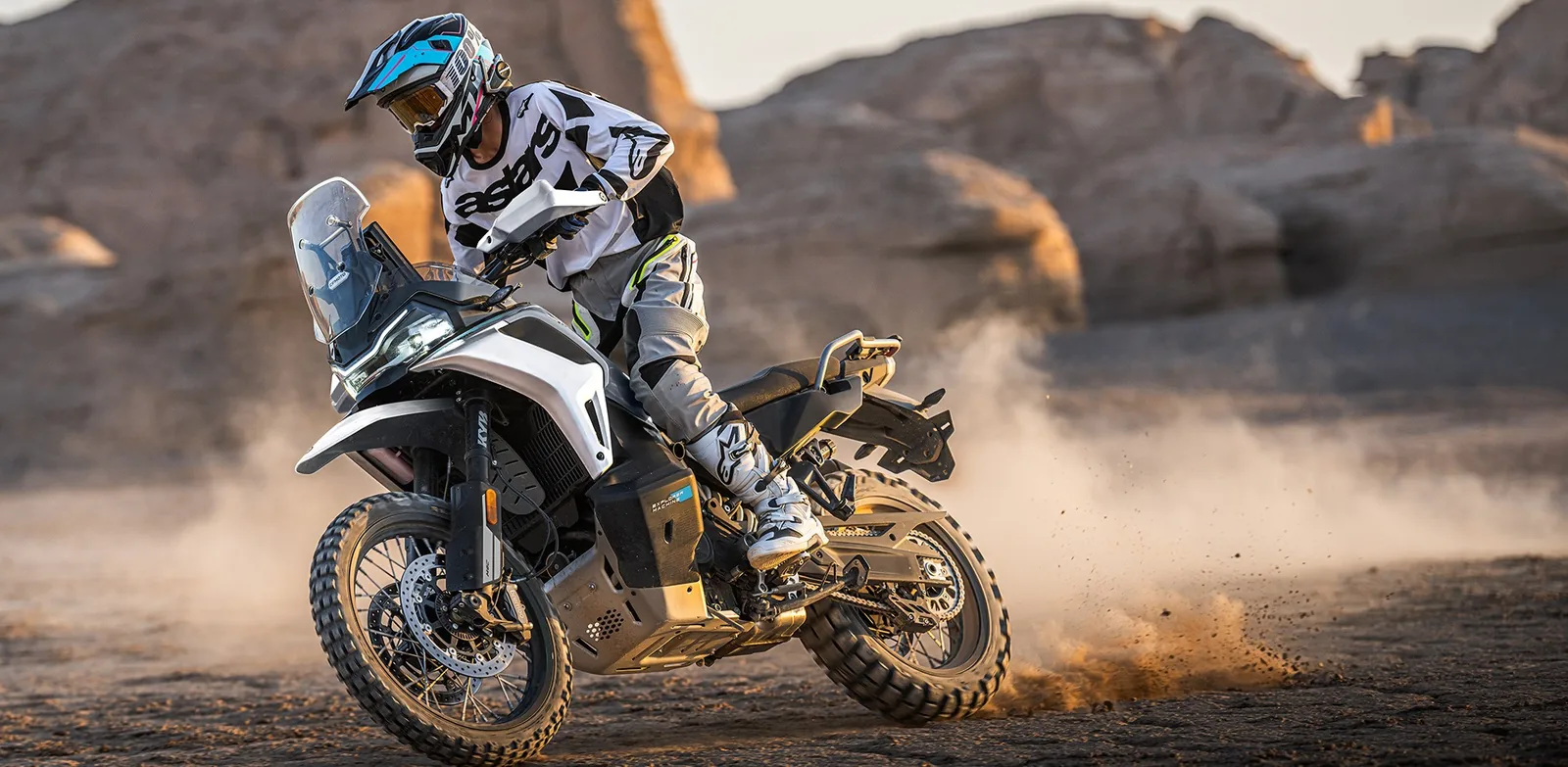 Rider sliding the rear wheel of a grey CFMoto 1000MT-X on hard-packed dirt among sandstone rocks, with dust coming off the back tyre
