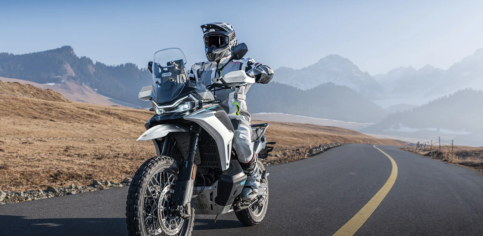 Rider on a grey CFMoto 1000MT-X on a curving tar road through dry grassland with snow-capped mountains behind, showing the adjustable windscreen and LED headlight