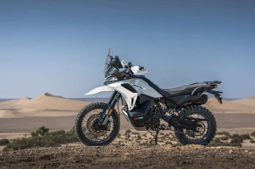 CFMoto 1000MT-X adventure bike in Aerolite Grey parked on its side stand on a stony rise with sand dunes behind, seen from the left side