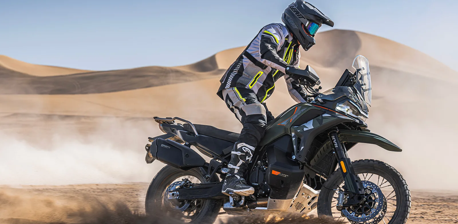 Rider standing on the pegs of a Tactical Green CFMoto 1000MT-X riding through sand with dunes behind, showing the low-slung fuel tank, bash plate and Brembo front brake