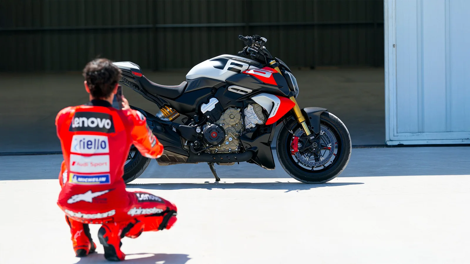 Francesco Bagnaia in Ducati Lenovo leathers crouching to photograph a Diavel V4 RS parked on its sidestand outside a hangar
