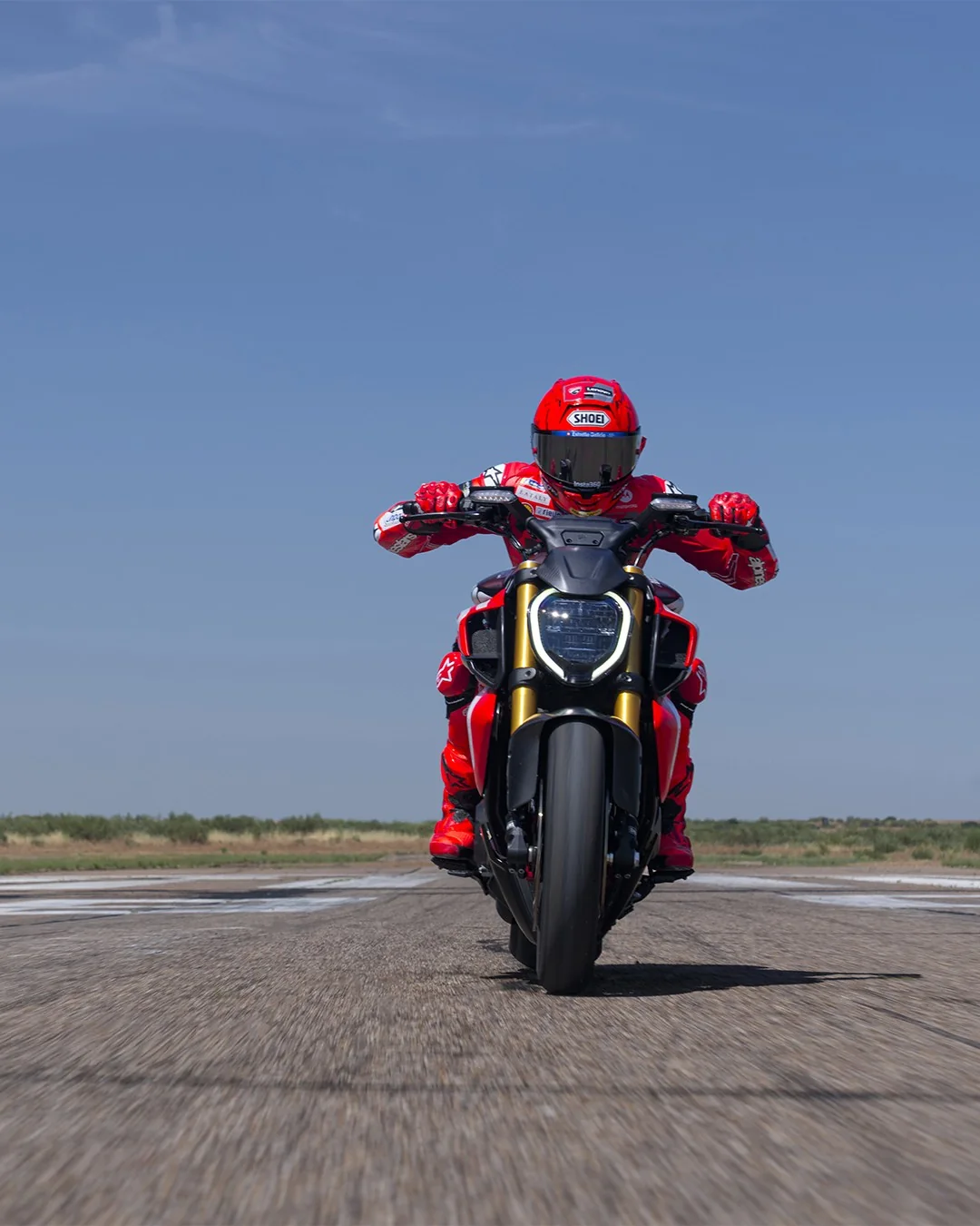 Head-on view of Marc Marquez riding a Ducati Diavel V4 RS towards the camera on an airstrip under a blue sky