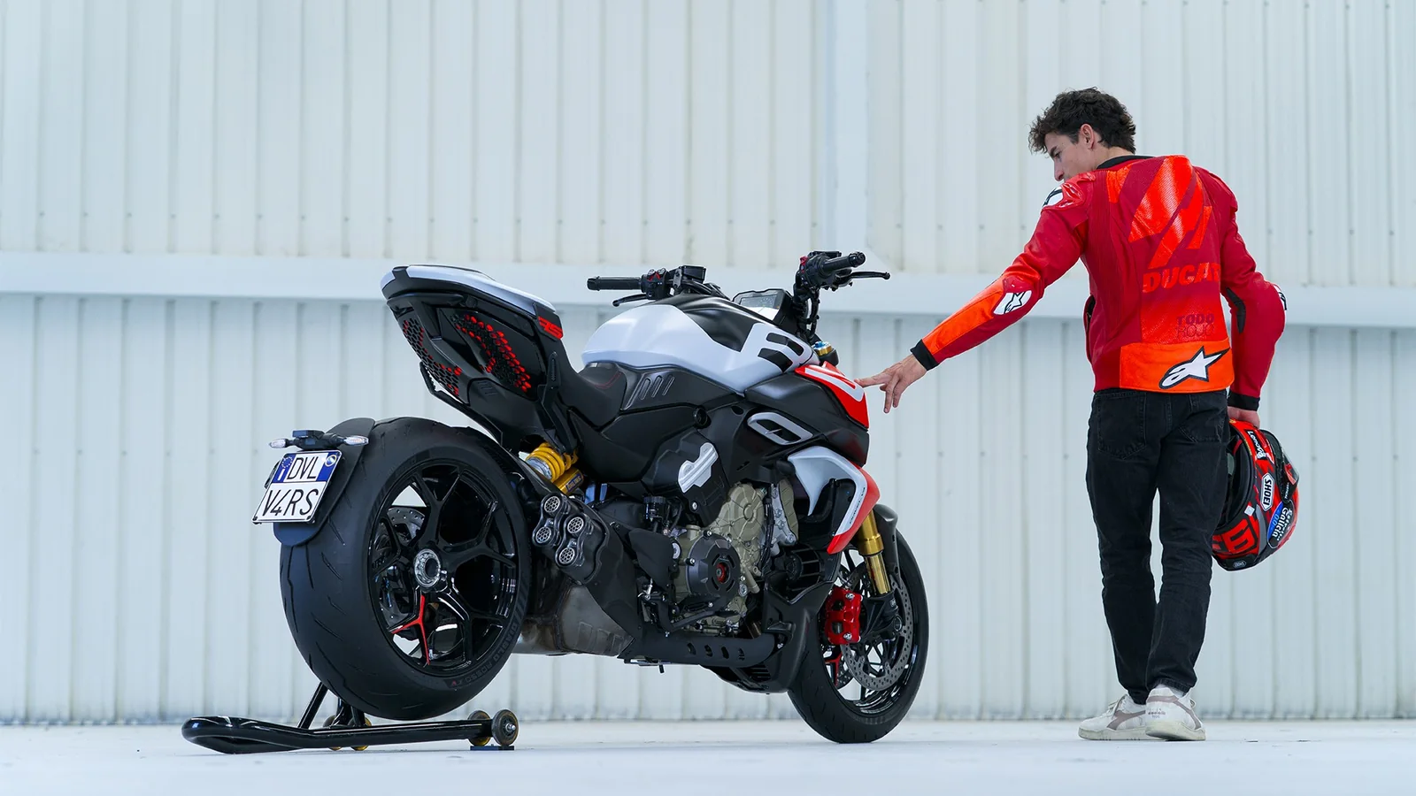 Marc Marquez in a red Ducati jacket touching the tank of a Diavel V4 RS on a paddock stand in a white hangar, seen from behind with the quad exhaust tips and 240 rear tyre visible