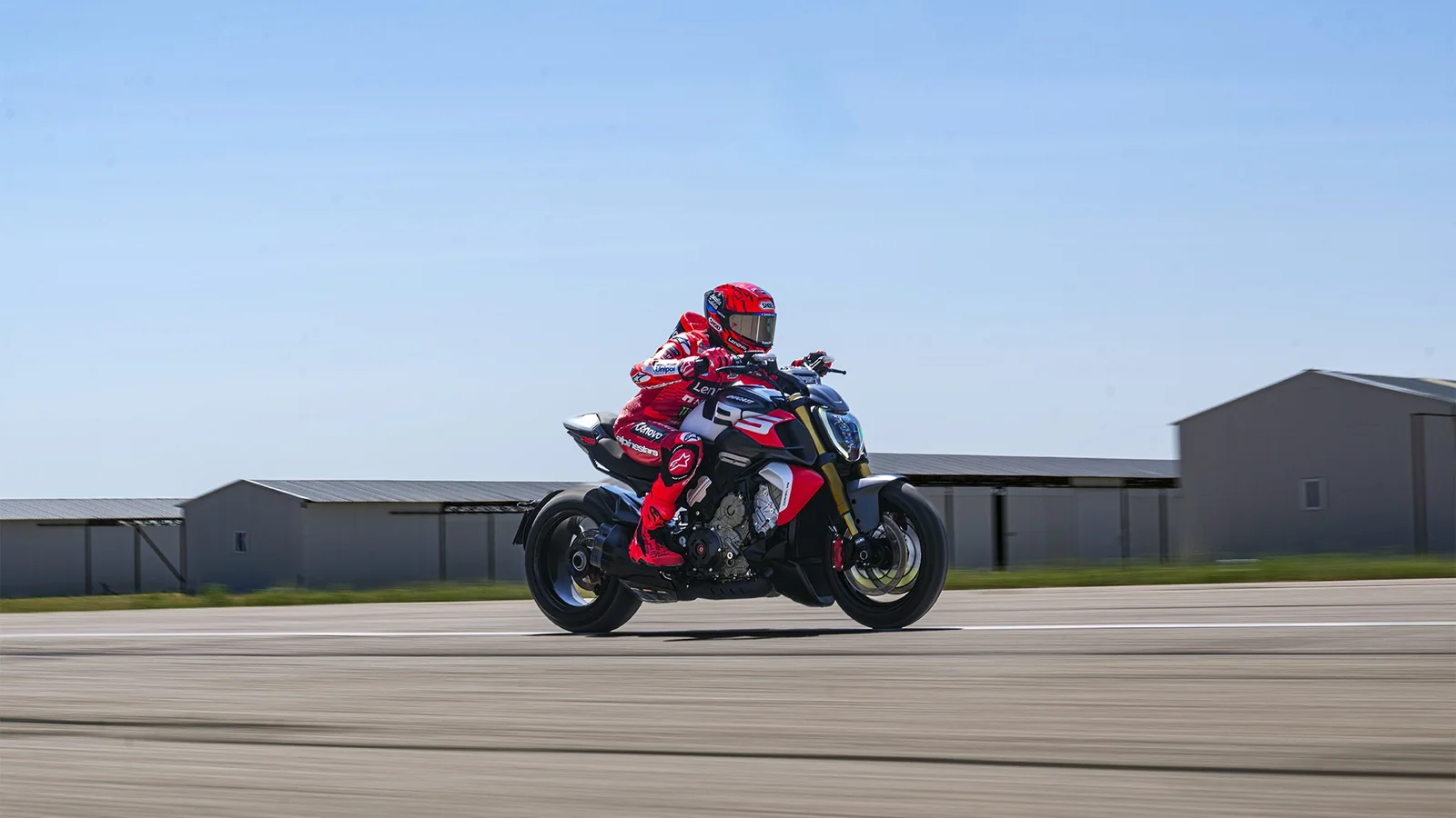Marc Marquez in Ducati Lenovo leathers accelerating a Diavel V4 RS along an airstrip with hangars behind