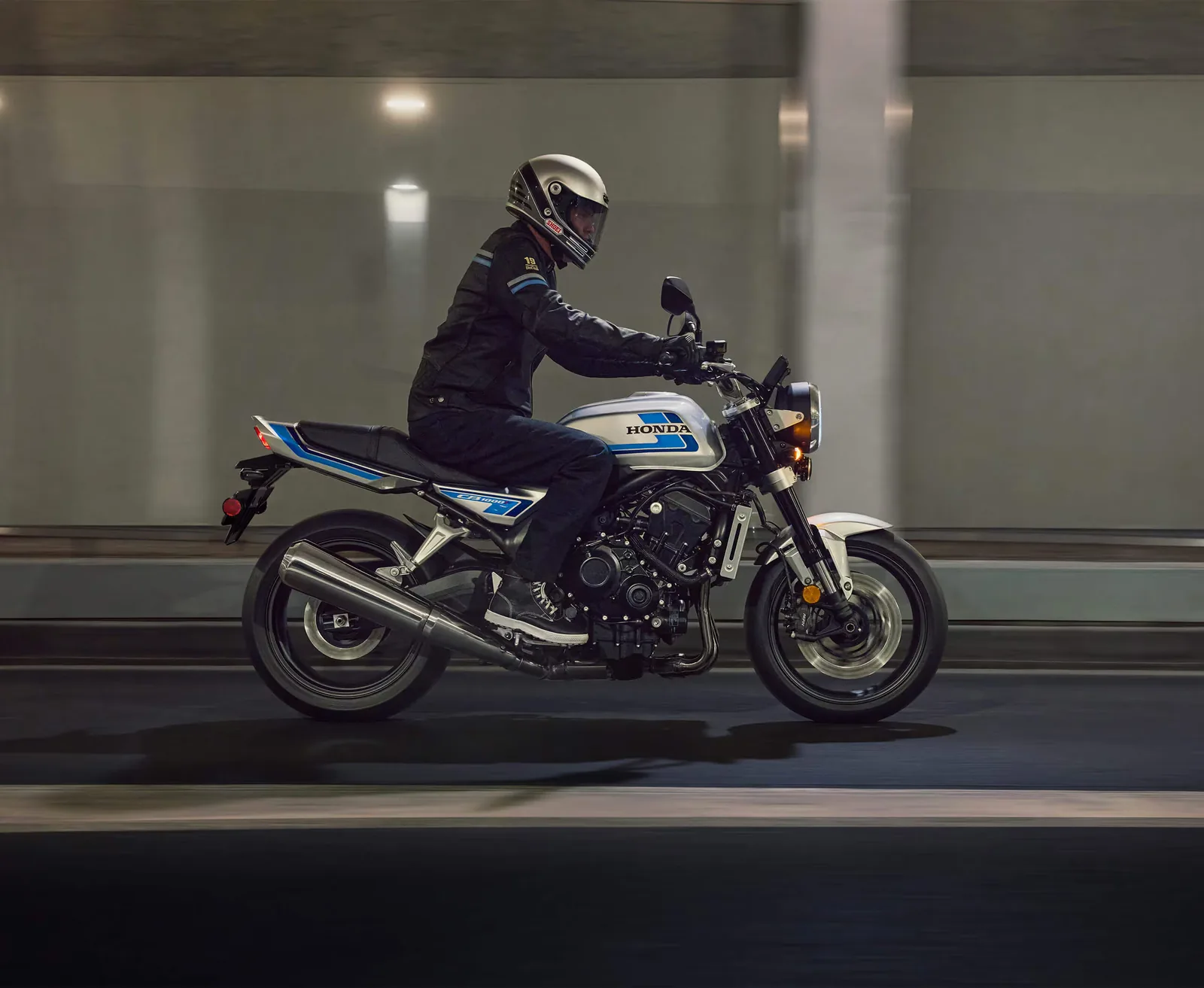 Rider in a black jacket and silver helmet on a 2026 Honda CB1000F, seen side-on riding through a concrete underpass