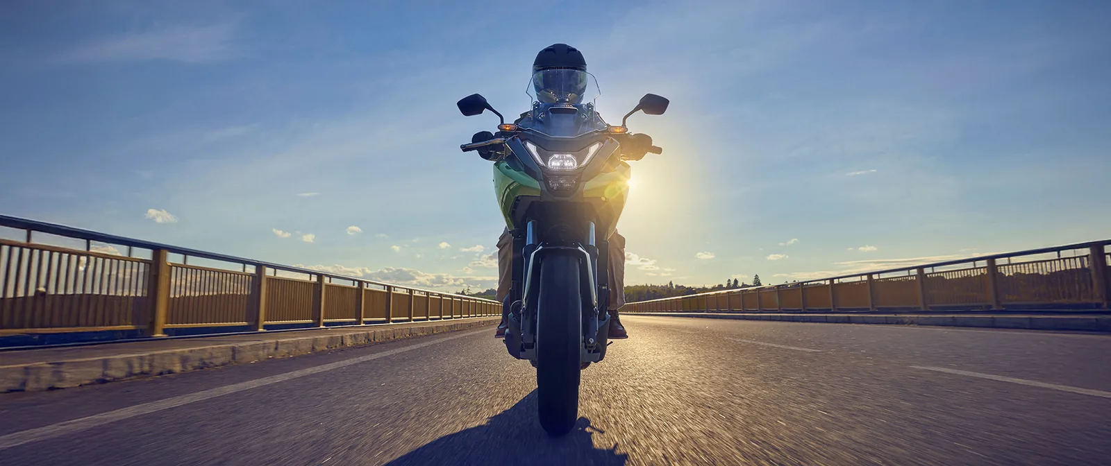 Head-on view of a rider on a green Honda NC750X crossing a bridge into low sun, showing the new LED headlight and mirrors