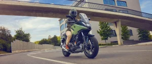 Rider in a green helmet and khaki trousers on an Earth Khaki Honda NC750X riding under a concrete footbridge past a modern building