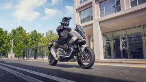 Rider in black adventure kit on a sand-coloured Honda X-ADV riding along a city street past a glass-fronted building and a bus shelter