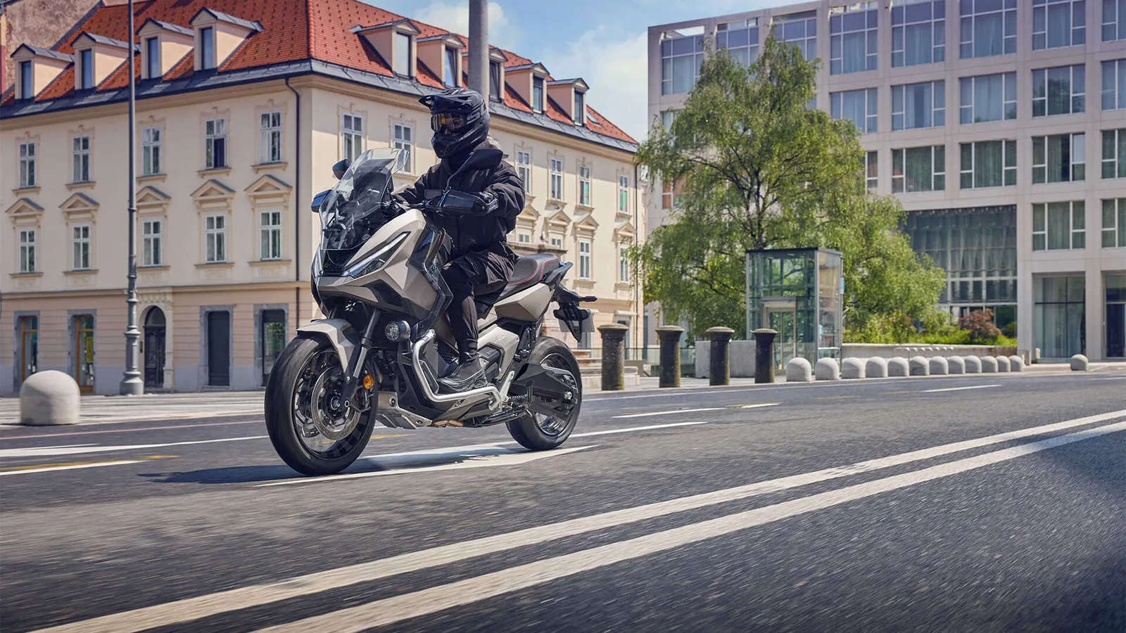 Rider on a grey Honda X-ADV with wire wheels and crash bars riding past old apartment buildings and a modern glass office on a city street