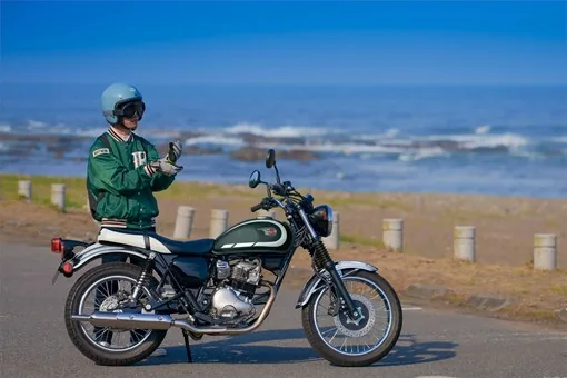 Rider in a green varsity jacket and open-face helmet standing beside a green Kawasaki W230 on a coastal road with the sea behind