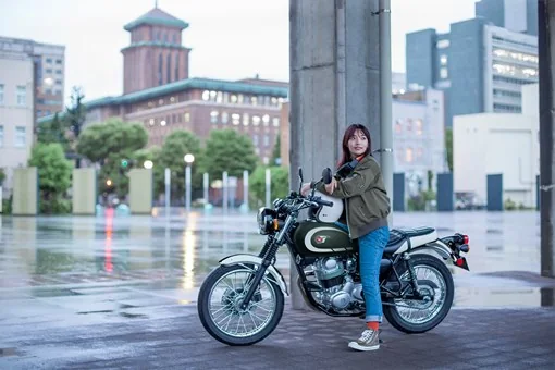 Woman in a bomber jacket and jeans standing with a green Kawasaki W230 under concrete pillars in a city square at dusk