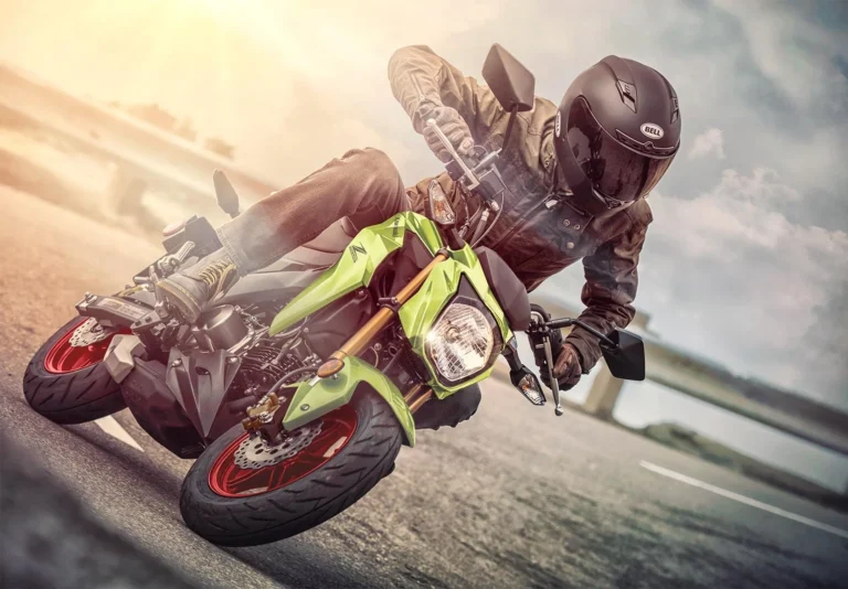 Rider in a black Bell helmet leaning a Metallic Yellowish Green Kawasaki Z125 Pro through a corner on a coastal road, red wheels and gold upside-down fork visible