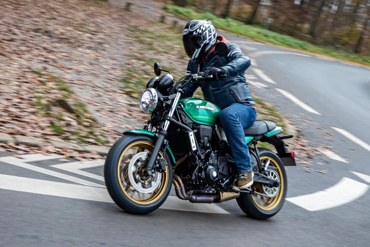 Rider in a leather jacket and jeans leaning a green Kawasaki Z650RS through a hairpin bend on a leaf-strewn road