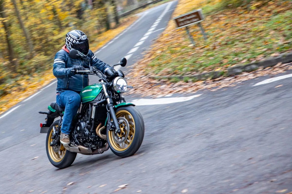 Rider on a green Kawasaki Z650RS riding uphill towards the camera on a narrow autumn road through woods