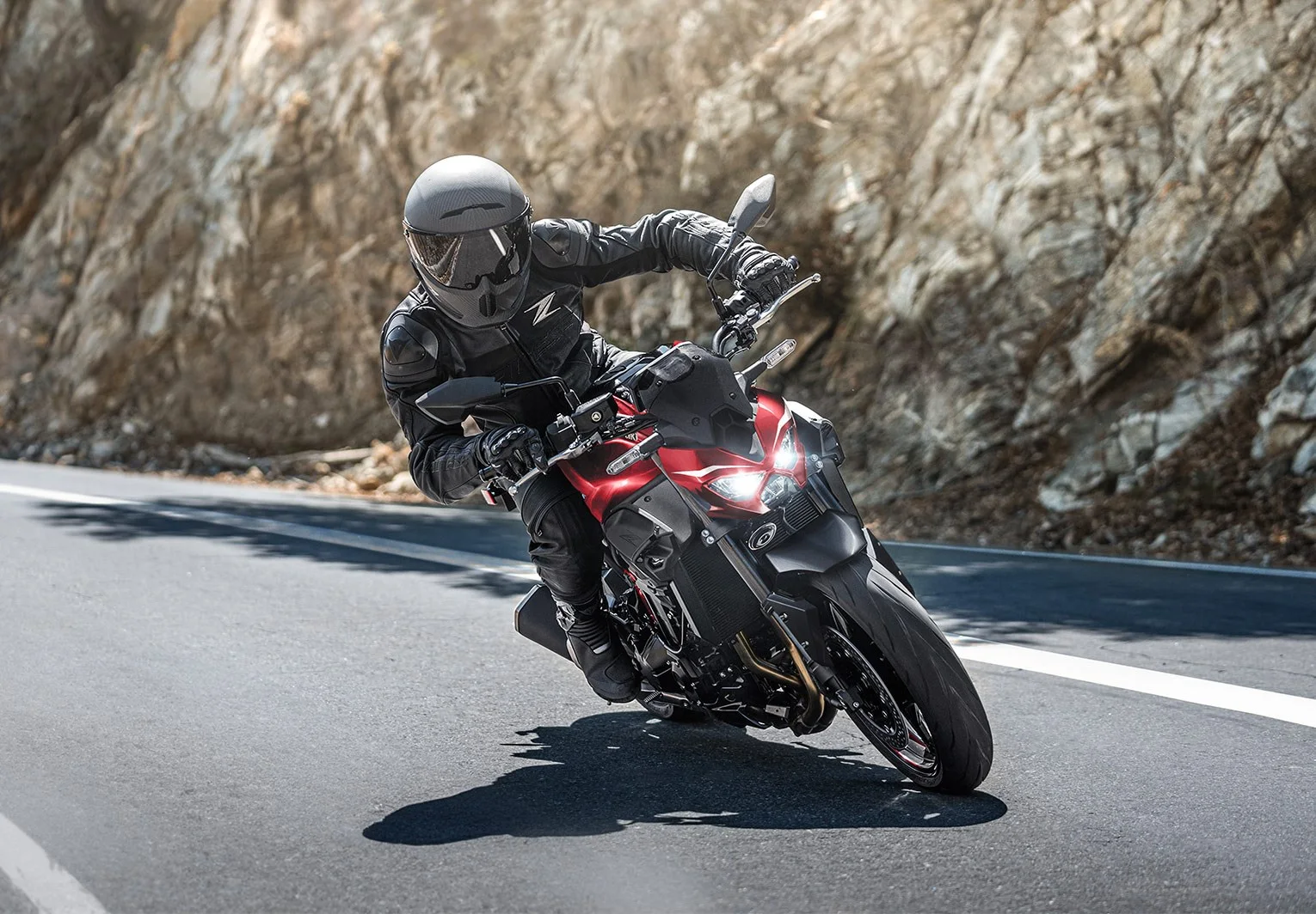 Rider leaning a red Kawasaki Z900 through a corner on a mountain road with a rock face behind