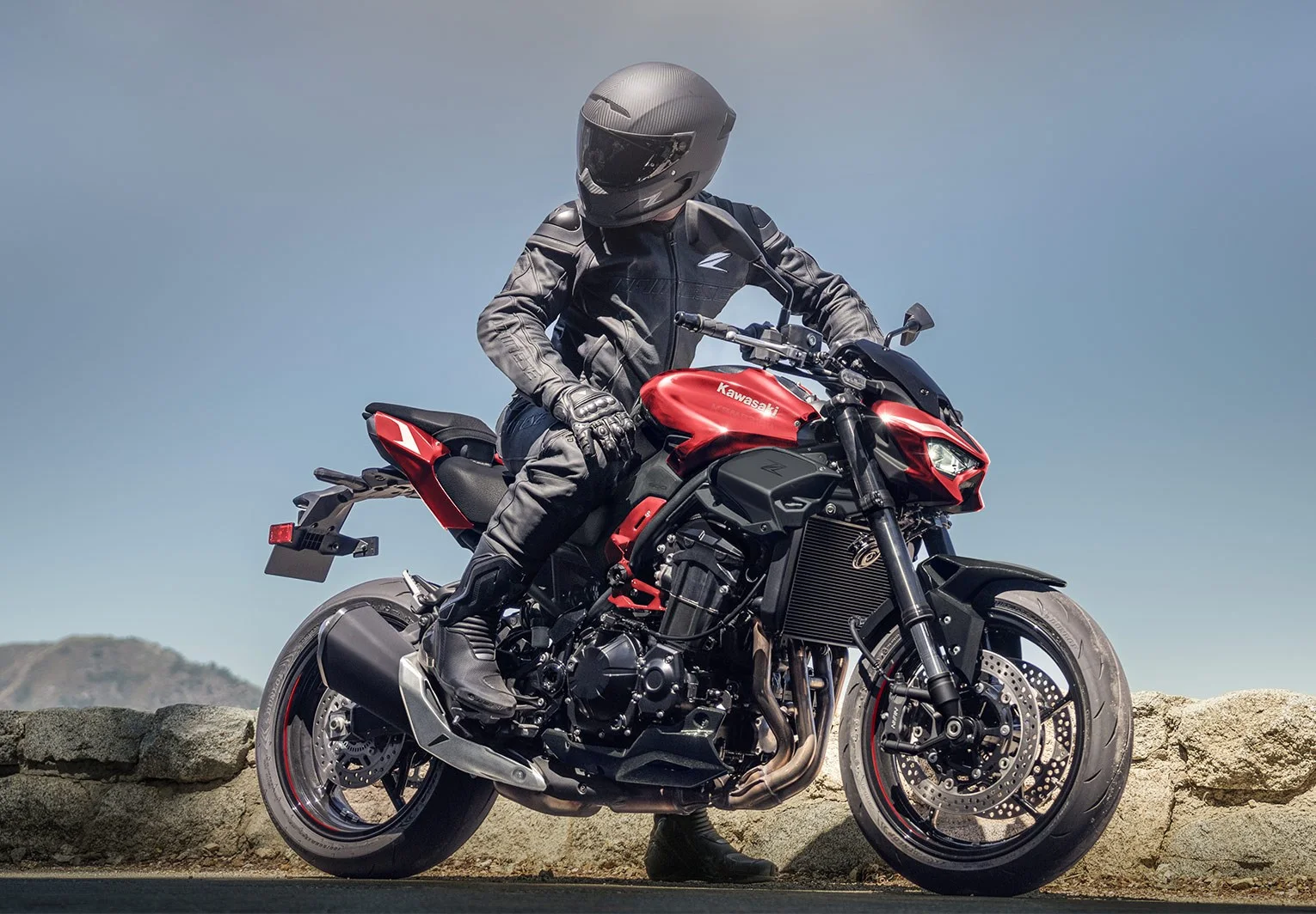 Rider in black leathers seated on a red Kawasaki Z900 beside a stone wall with mountains behind