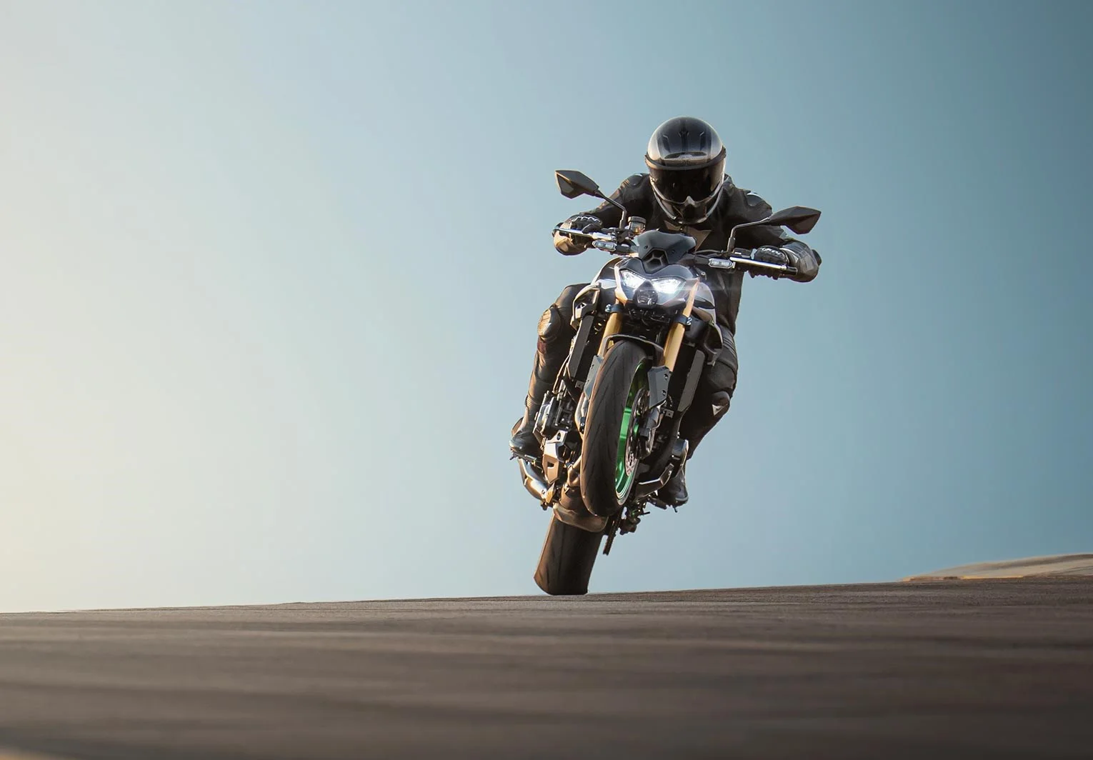 Rider in black leathers lifting the front wheel of a Kawasaki Z900 on a road, headlight lit, green rim visible