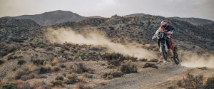Rider in KTM orange kit and a Red Bull helmet jumping a white and orange KTM 1390 Super Adventure R over a crest on a desert trail with dust and mountains behind