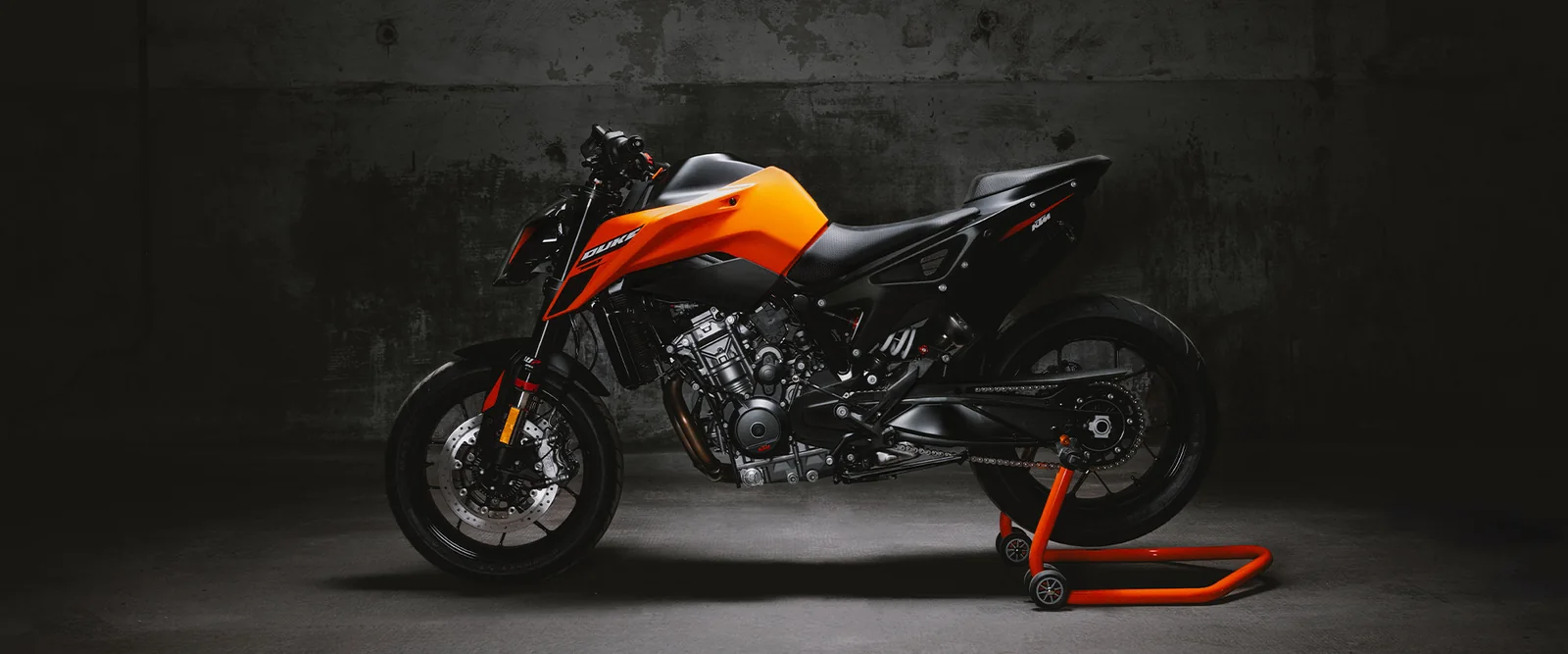 Orange KTM 790 Duke on a rear paddock stand against a concrete wall, seen side-on, showing the short tail and exposed twin-cylinder engine