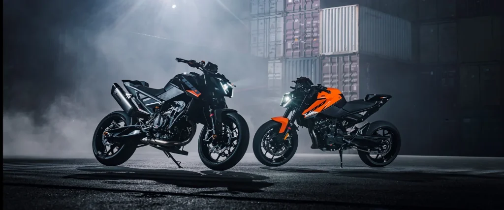 Two KTM 790 Duke motorcycles parked in a container yard at night, one black with accessory parts and one in orange, lit from behind through haze