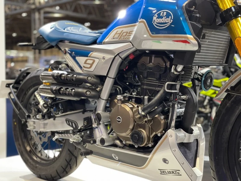 Three-quarter view of the Mondial HPS 125 Ubbiali liquid-cooled twin-cam single with bronze clutch cover, radiator, twin exhausts and Dell'Orto-branded bash plate on a show stand