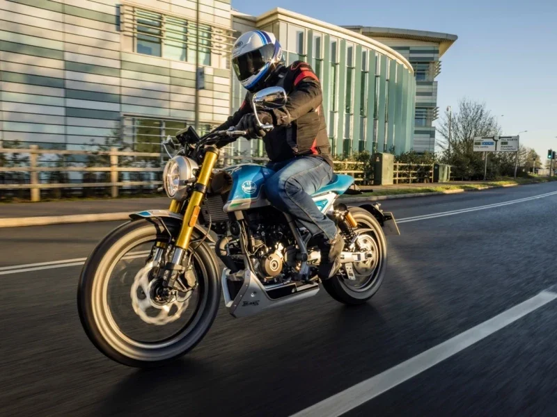 Front three-quarter view of a rider on a Mondial HPS 125 Ubbiali Limited at speed on a road past a glass office building, round headlight and gold forks visible
