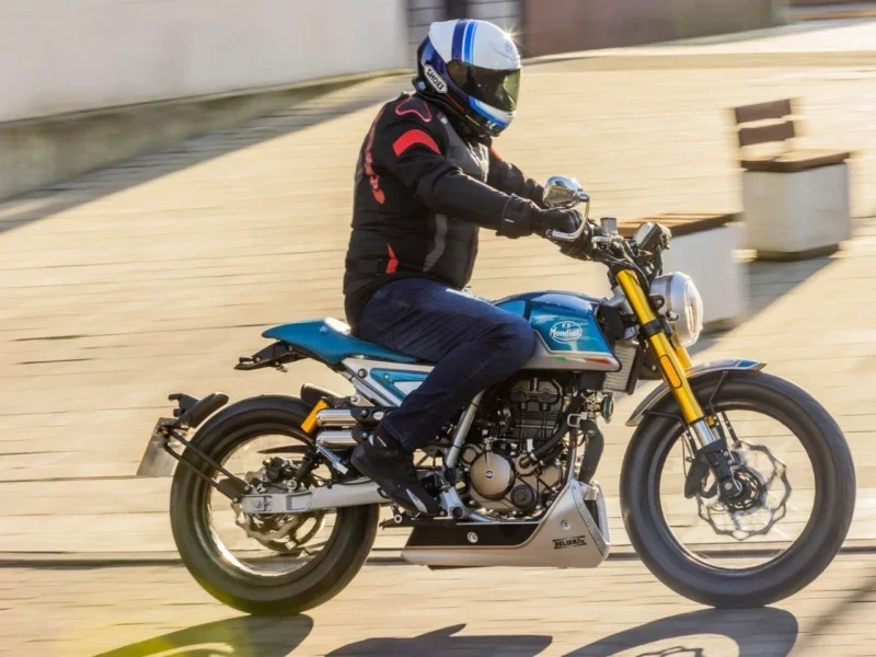 Rider in a white and blue helmet riding a light blue Mondial HPS 125 Ubbiali past a paved square, showing the spoked wheels, bash plate and quilted seat