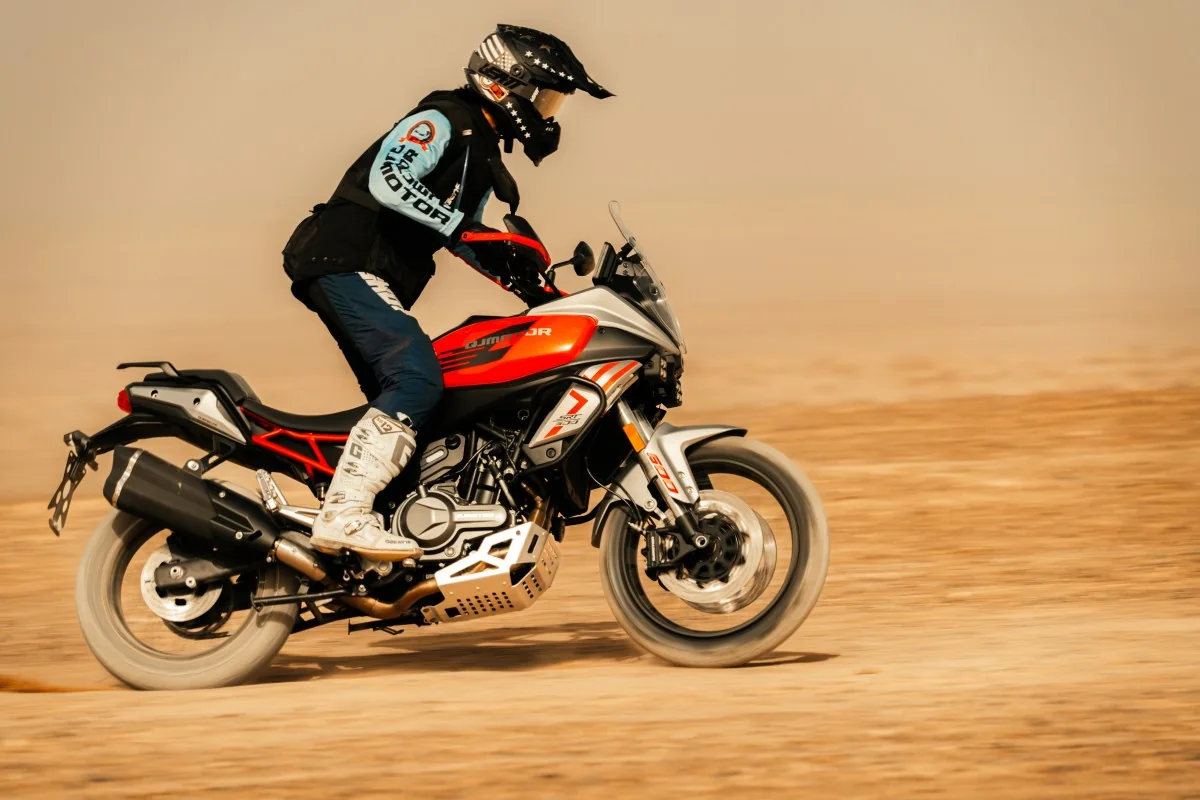 Rider standing on the pegs of a red QJMotor SRT 600 at speed on a dusty gravel plain