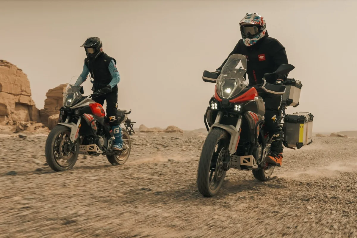 Two QJMotor SRT 600 adventure bikes ridden on a stony desert track, one carrying aluminium panniers and a top box