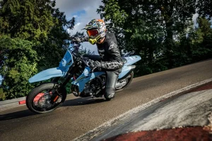 Rider in black leathers cornering a Sky Gray Suzuki DR-Z4SM supermoto hard on a kart track, knee close to the tar, trees behind