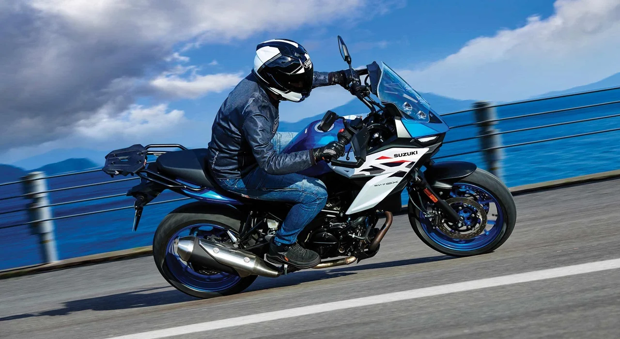 Rider in a white helmet and denim jacket cornering a 2027 Suzuki SV-7GX on a coastal road, seen from the right with the exhaust, trellis frame and knuckle covers visible