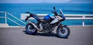 2027 Suzuki SV-7GX in Pearl Brilliant White and Metallic Triton Blue parked side-on beside a guard rail with the sea behind, showing the half fairing, adjustable screen, blue wheels and rear carrier