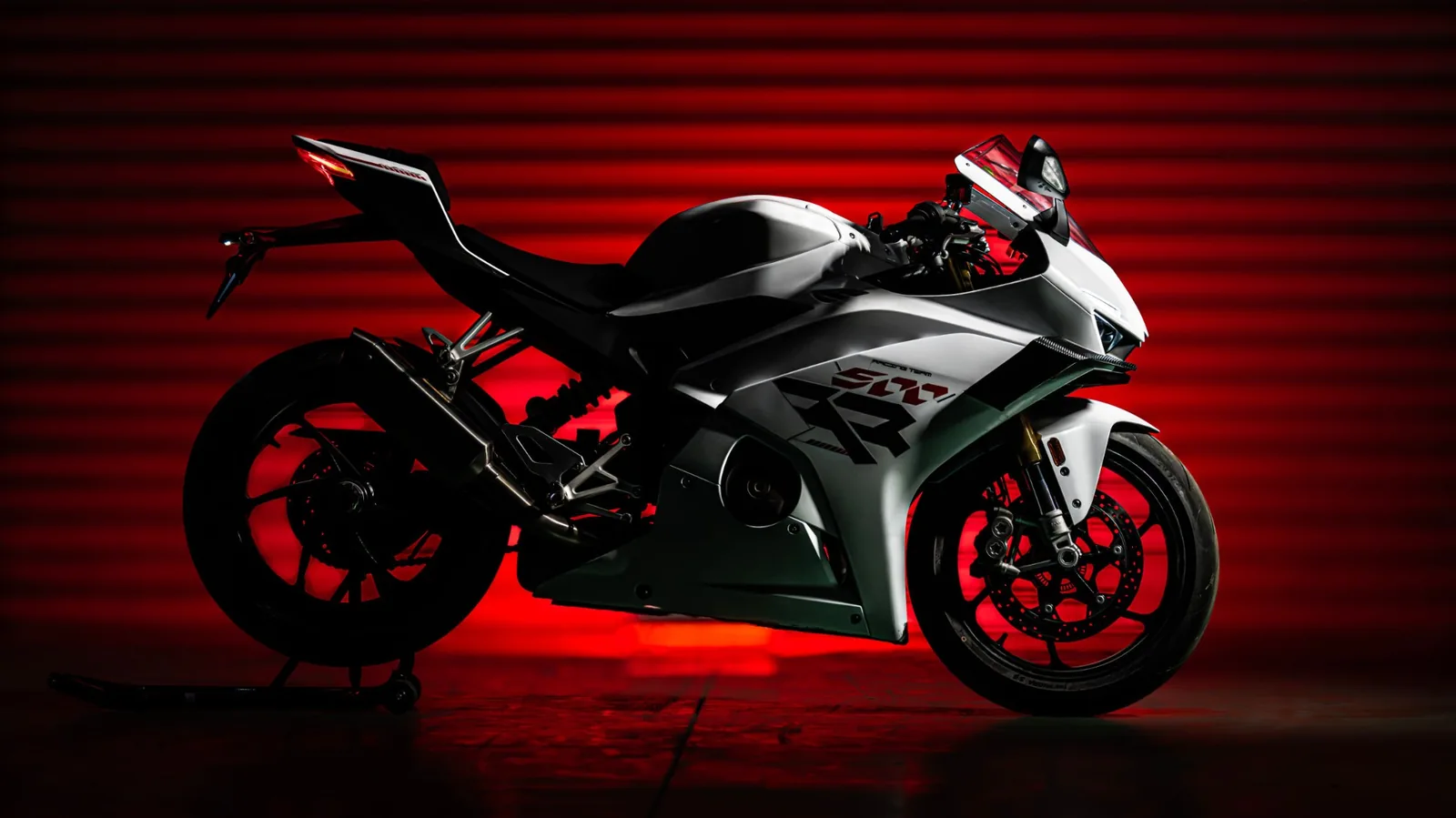 ZXMOTO 500RR sportsbike in white on a paddock stand in a dark studio lit red from behind, showing the single-sided swingarm and winglets